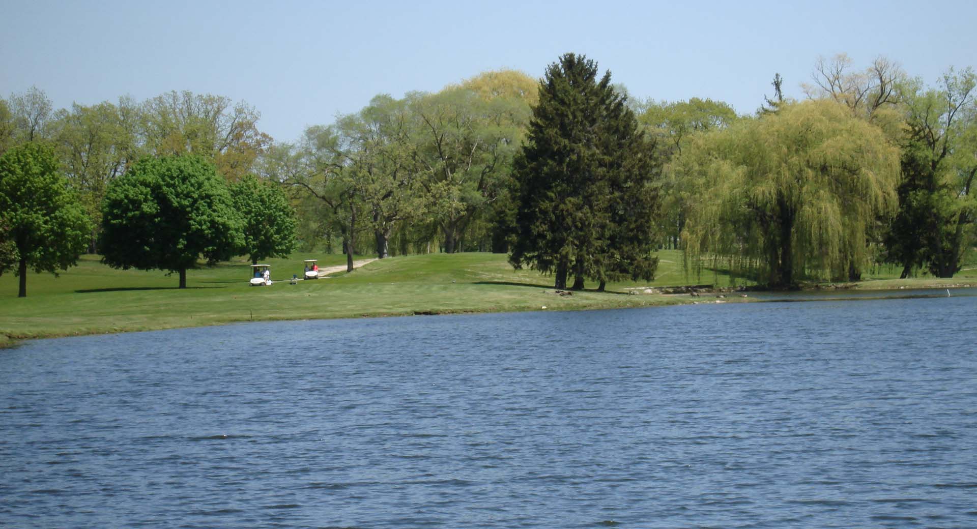 Course greens with lake in the foreground and trees in the background with a cart path used by golfers on the right